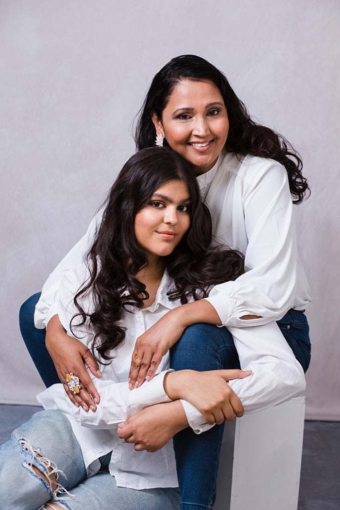 A warm and stylish mother daughter photo shoot capturing the close bond between a mother and her teenage daughter, in Asian Indian ethnicity. Both are dressed in coordinated white blouses and jeans, sitting closely together with genuine smiles. The portrait emphasizes love, connection, and a relaxed, modern style, photographed in a North London studio.