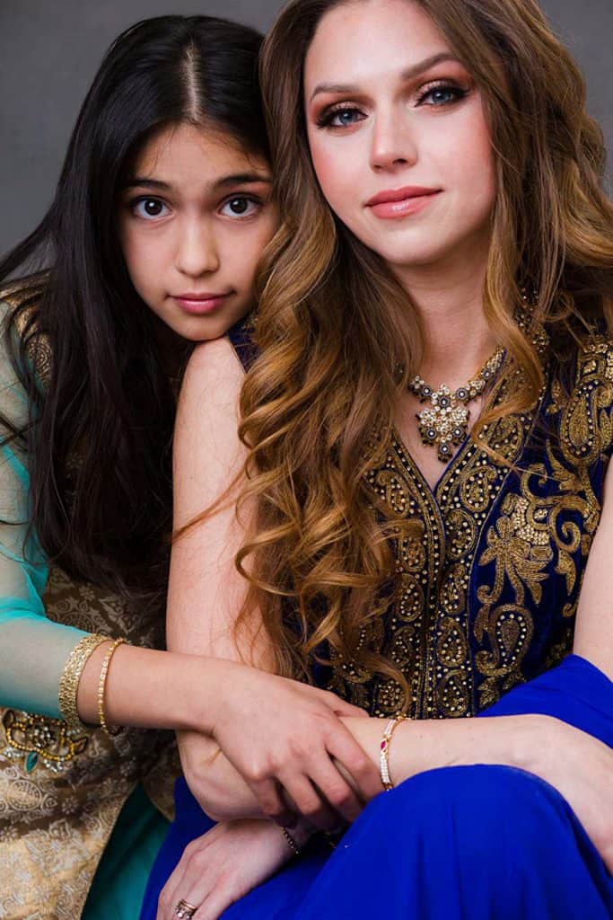 Mother and daughter in an elegant makeover photoshoot wearing beautiful traditional attire. The mother is dressed in a royal blue embellished sari, with glamorous makeup and jewelry, while her daughter leans on her, wearing a light blue Indian national dress. The portrait captures the close bond and beauty of their relationship, showcasing heritage and a luxurious experience.