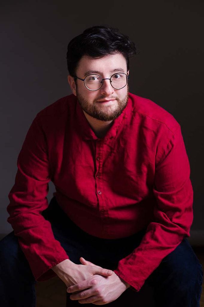 Personal-brand-photography-man-in-red-shirt-glasses-sitting-leaning-forward-dark-background