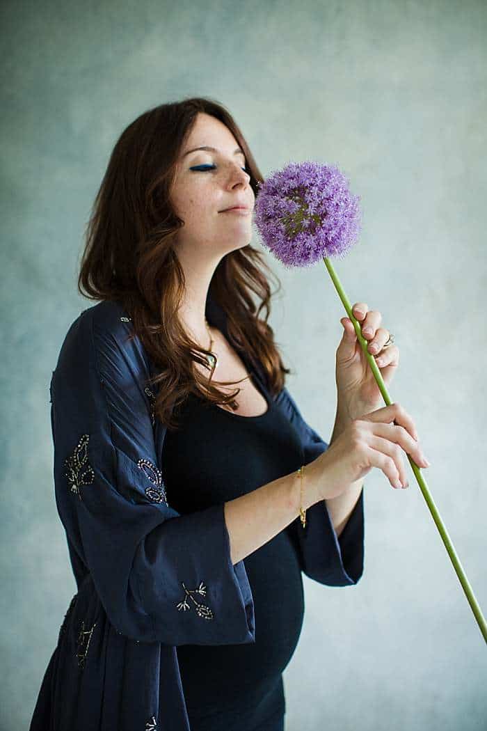 brunette-pregnant-woman-in-blue-robe-sensually-smelling-purple-flowr-stem-light-blue-background-in-maternity-photography-london-studio