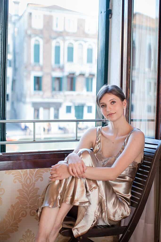 portrait-photoshoot-in-venice-hotel-room-girl-sitting-on-chair-by-window-venetian-houses-in-background
