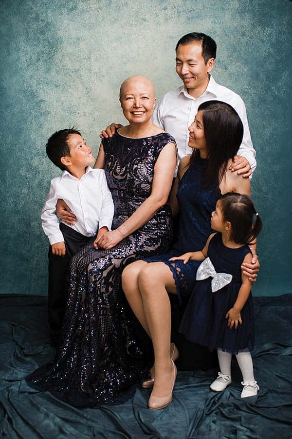A heartwarming three generation family portrait taken in a professional photography studio in London. The image features a beautifully dressed family of five, with a mother in an elegant navy blue gown, surrounded by her son, daughter in law and grand children, all dressed in coordinated outfits. The mother has undergone cancer treatment and hence the head is temporarily bald. The setting has a soft, artistic blue-green backdrop, creating a timeless and emotional moment captured in a professional family photoshoot.