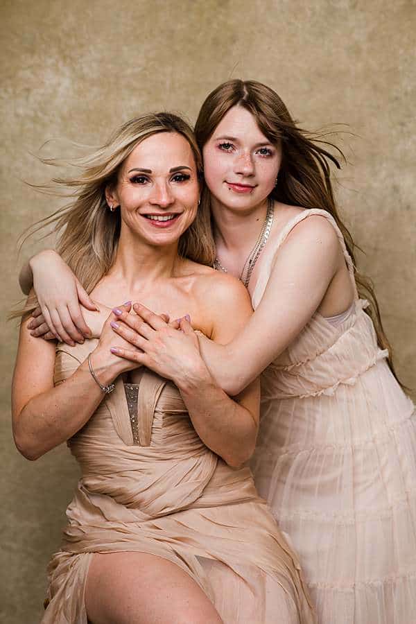 Glamourous mother daughter photoshoot portrait in beige dresses embracing and smiling in a studio portrait with a neutral background.